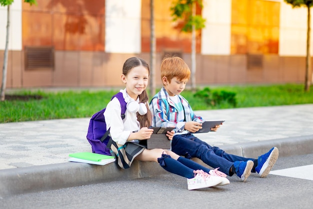 A boy and a girl with briefcases or backpacks are sitting at the school and reading a book and tablet going back to school brother and sister are studying doing homework on the street