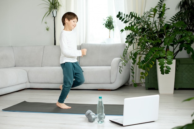 The boy goes in for sports at home online. The child does exercises in the room.