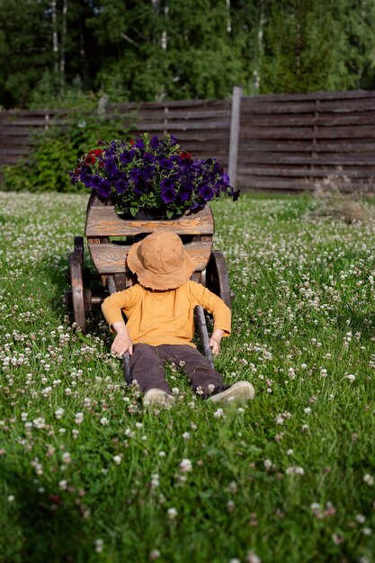 A boy in a hat is sitting on the grass near a decorative cart with flowers