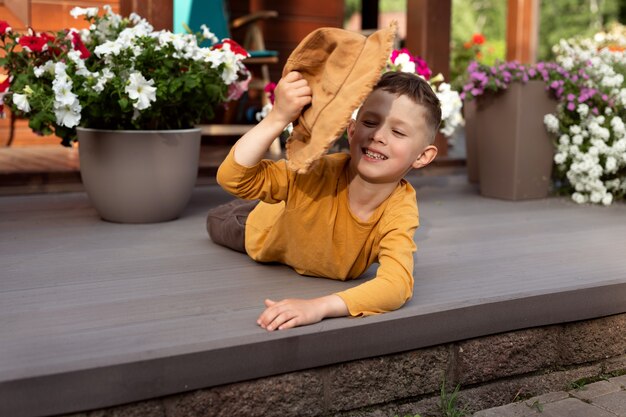 A boy in a hat lies on the veranda of a wooden house surrounded by flowers