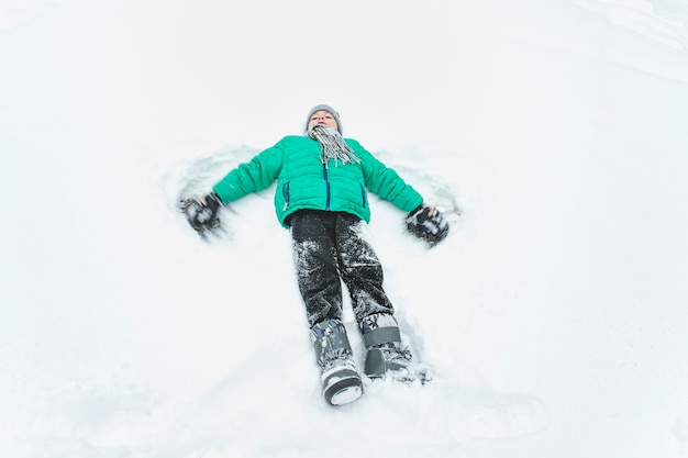 Boy in hat scarf and green jacket lying in snow and making angel Portrait Closeup Winter day