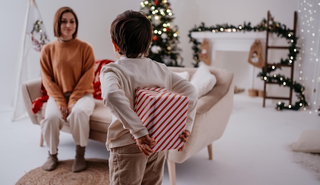 Boy hiding present behind his back and preparing giving it to his mother while celebrating Christmas