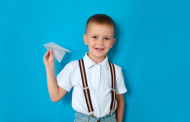 Boy holding a paper airplane on a blue background A photo of a young excited schoolboy with a happy positive smile throws an airplane model into the air