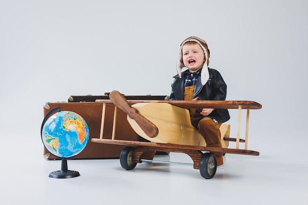 The boy is crying in a leather jacket and a pilot's hat a wooden plane brown suitcases Children's wooden toys Baby emotions