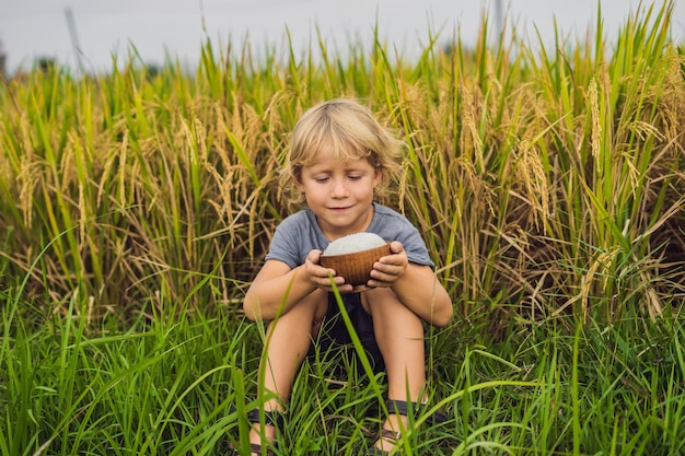 Boy is holding a cup of boiled rice in a wooden cup on the background of a ripe rice field. Food for children concept