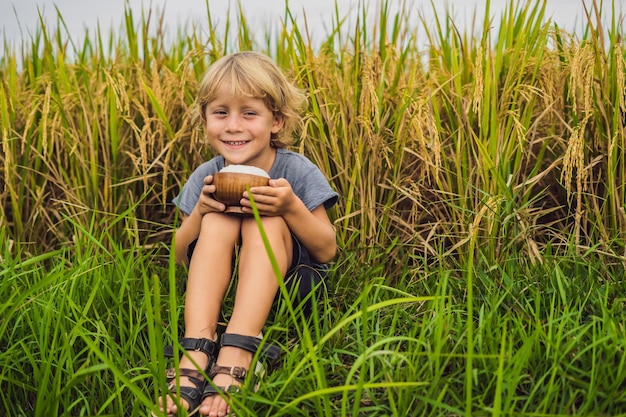 Boy is holding a cup of boiled rice in a wooden cup on the background of a ripe rice field. Food for children concept.