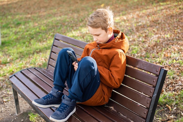 A boy is playing on his phone while sitting on a park bench