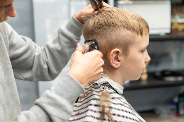 A boy is sitting in a barber shop cutting his hair with a clipper