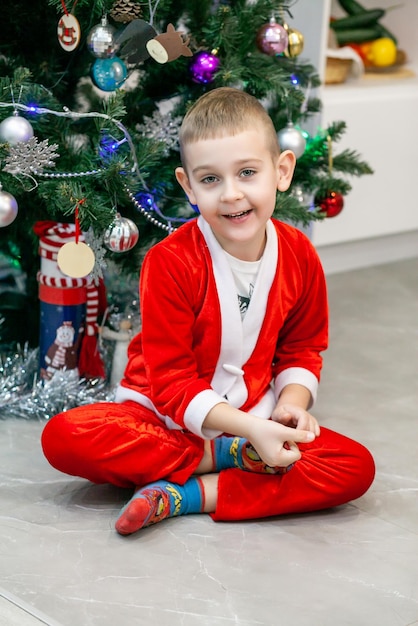 The boy is sitting under the Christmas tree in a Santa costume without a hat looking at the camera and smiling