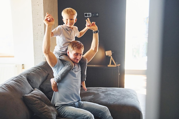 Photo boy is sitting on the man's shoulders father and son is indoors at home together