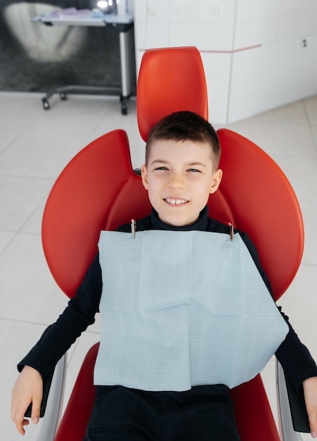 The boy is sitting in a red dental chair and smiling in modern white dentistry Treatment and prevention of caries since childhood Modern dentistry and prosthetics