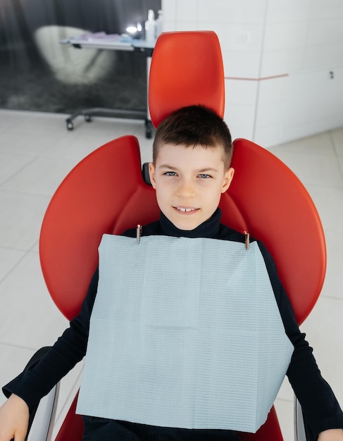 The boy is sitting in a red dental chair and smiling in modern white dentistry Treatment and prevention of caries since childhood Modern dentistry and prosthetics