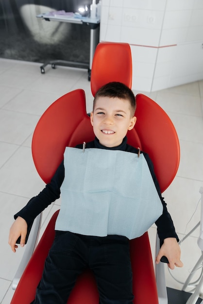 The boy is sitting in a red dental chair and smiling in modern white dentistry Treatment and prevention of caries since childhood Modern dentistry and prosthetics