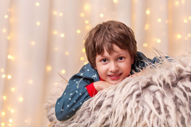 Boy is smiling watching the Christmas tree in lights and window with garland surrounds holiday atmosphere