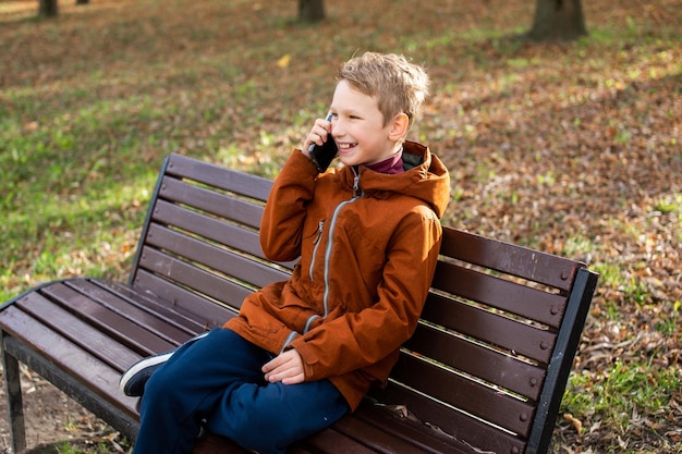 A boy is talking on the phone and laughing while sitting on a park bench in autumn