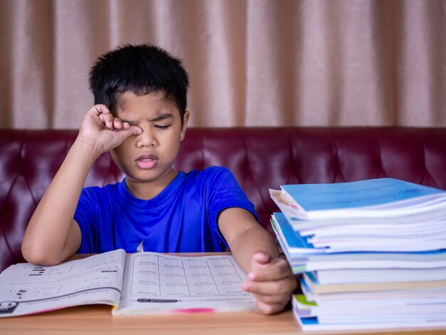 A boy is tired of reading a book on a wooden table. with a pile of books beside The background is a red sofa and cream curtains.