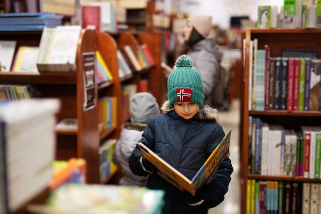 Boy in jacket and hat reaching a book from bookshelf at the library Learning and education of european kids