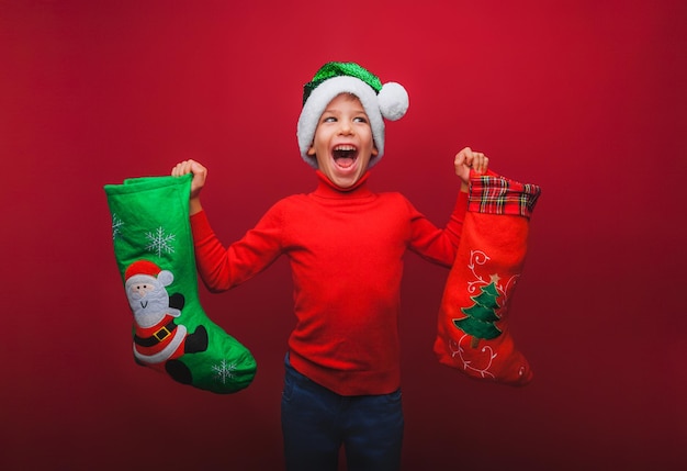 A boy in a knitted red sweater and a Santa Claus hat holds a Christmas boot for gifts Cute baby is waiting for Christmas and a new kind insulation on a red background