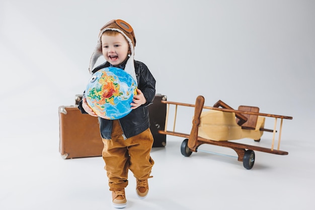 A boy in a leather jacket and a pilot's hat a wooden plane a globe brown suitcases Children's wooden toys