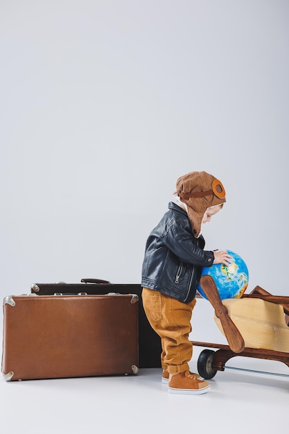 A boy in a leather jacket and a pilot's hat a wooden plane a globe brown suitcases Children's wooden toys