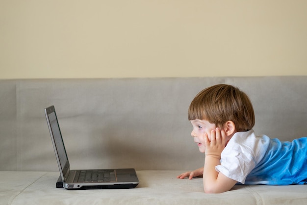 The boy lies on a bright sofa and looks at a laptop.