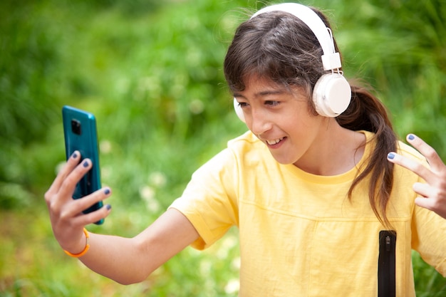 Boy listening to music from his mobile phone with white headphones and taking selfies next to a green meadow in spring