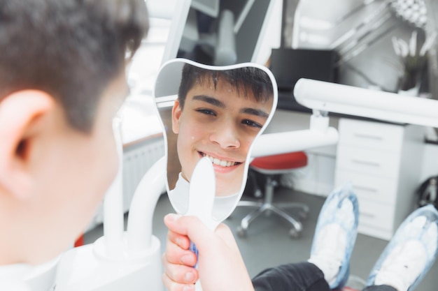 Boy looks in the mirror with a toothy smile sitting on a chair with a dentist in the dental office