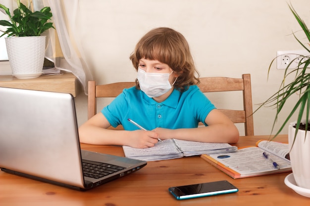 Boy in medical mask on her face doing homework and watching webinar at laptop.  Distance education, homeschooling, E-learning at home during quarantine concept