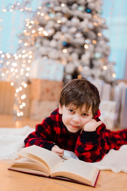 A boy in pajamas reads a book under a Christmas tree . New Year's mood. Reading books . Children's books.