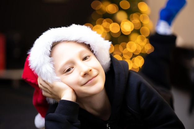 A boy in a red fluffy Santa fur hat on the background of Christmas tree lights