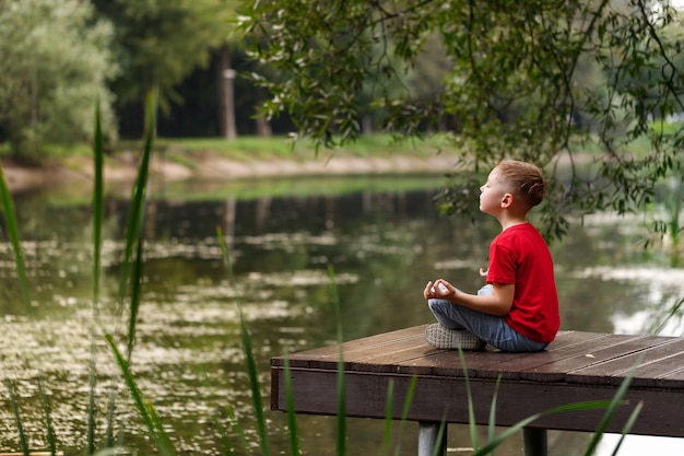 Boy in a red Tshirt meditates in the park Child in a yoga pose by the water