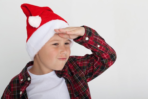 A boy in a red Tshirt and a Santa Claus hat is smiling Funny kid in the studio for Christmas on a white background The European kid smiles and makes a face New Year