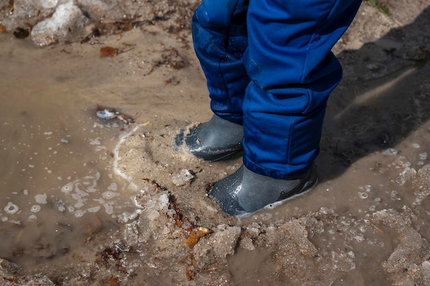 Boy's feet in rubber boots in muddy puddle with sand and snow Child boy of 67 year old walking and playingin wellies in melted earth Spring came