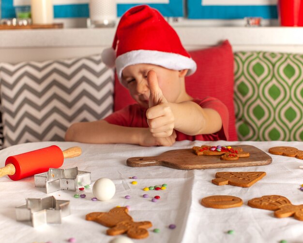 A boy in a santa claus hat holds a thumbs up surrounded by christmas cookies