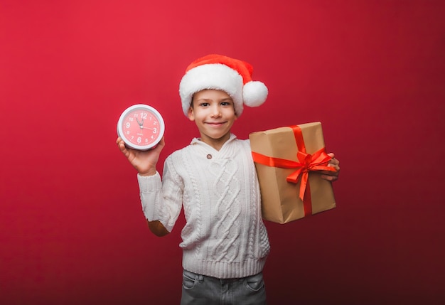 A boy in a Santa Claus hat holds a watch on a red studio background the concept of Christmas and new year a child in knitted clothes