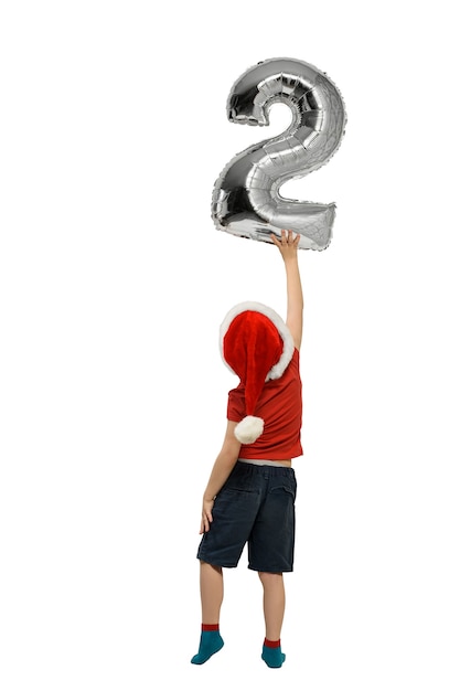 Boy in Santa hat holds up silver inflatable digit two. Back view. Isolated on white background.