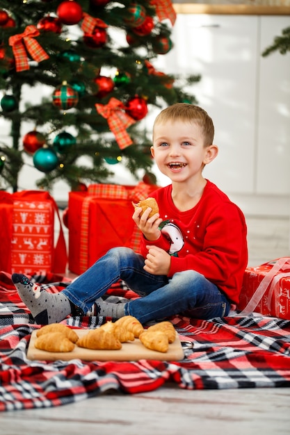 The boy sits near the Christmas tree in the Christmas kitchen. Holds a croissant in his hands. Cozy winter evenings at home. There are many Christmas gifts under the tree