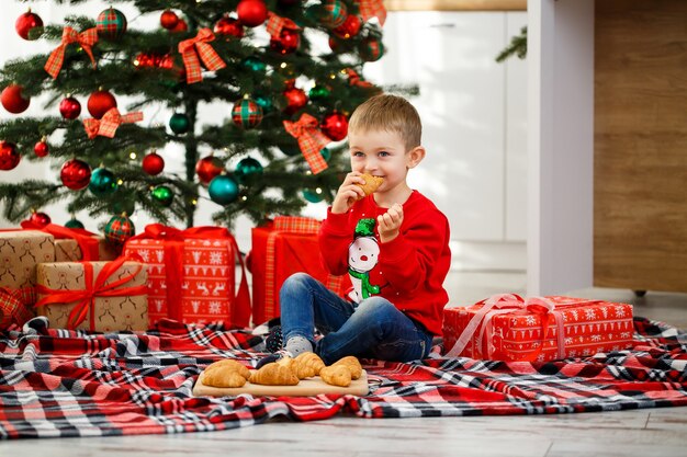 The boy sits near the Christmas tree in the Christmas kitchen. Holds a croissant in his hands. Cozy winter evenings at home. There are many Christmas gifts under the tree