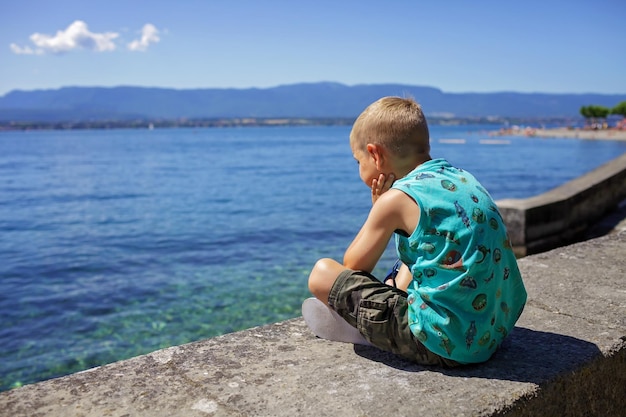 Boy sits on the quay of geneva lake and looks at water and mountains vacation and summer travel