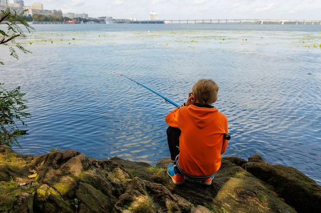 The boy sits with his back and catches fish with a fishing rod in the river