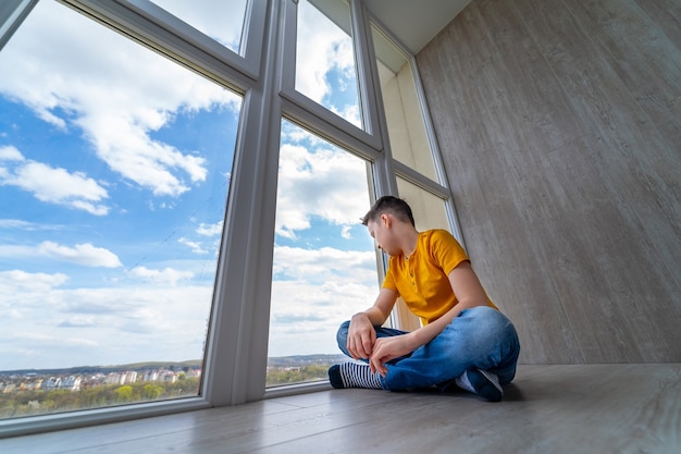 Boy sitting at floor at balcony. Kid looking at nature outside window. Isolation at home while quarrantine. Sad boy dreaming.