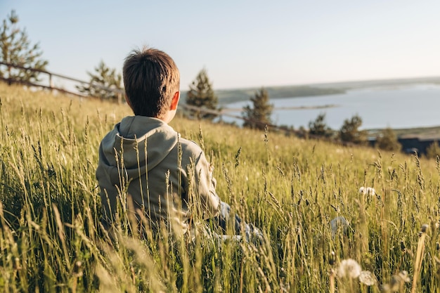 Boy sitting on top of hill in grass field and enjoying beautiful landscape view Rear view of teenager hiker resting in nature Active lifestyle Concept of local travel