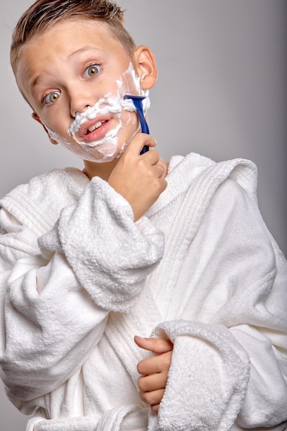 Boy son with pleasant appearance have shaving foam on face hold razor and going to shave stand in frot of mirror isolated on gray background young kid imitates father shocked facial expression