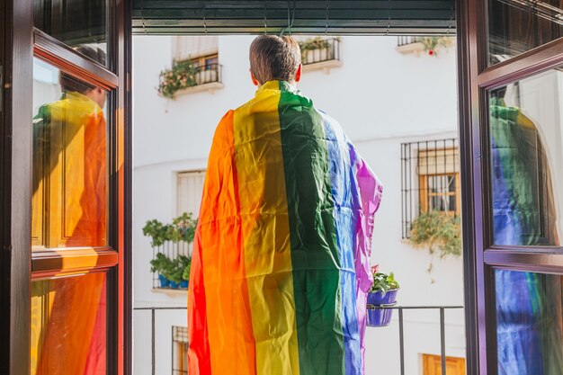 Boy standing on a balcony facing the street with a rainbow flag on his shoulders. lgtbi, gay