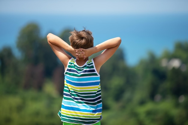 The boy stands with his hands folded in front of his head and admires the beautiful landscape