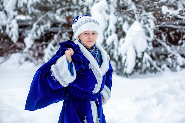 A boy in a suit of Father Frost with bag of gifts against the of snowy trees. Winter day