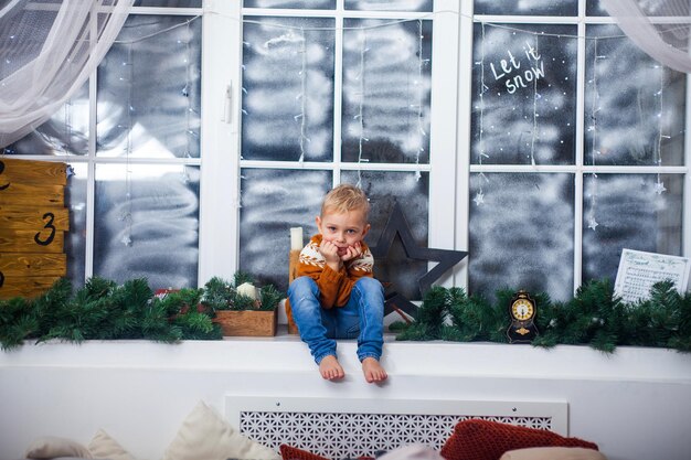 Boy in a sweater sits on windowsill at home waiting for miracles gifts and Christmas