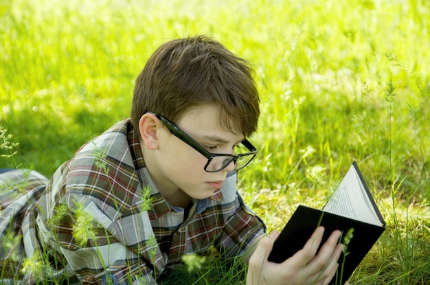 Boy teen with glasses lying in the green grass and reading a book Outdoor Park vacation