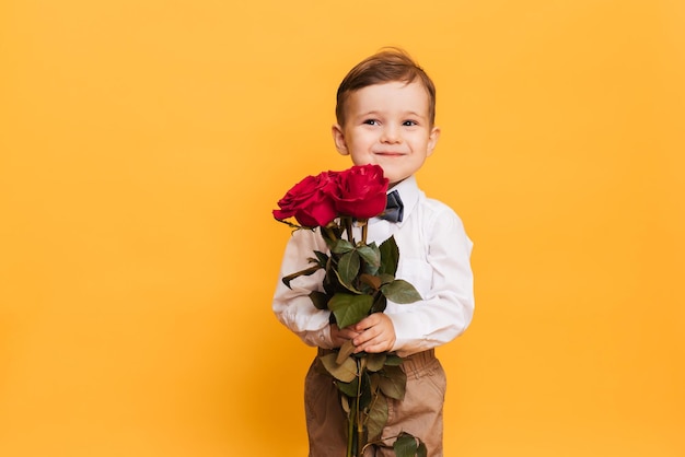 Boy in a white shirt pants and a bow tie on a yellow background holds in his hands bouquet a red roses A gift for my mother grandmother