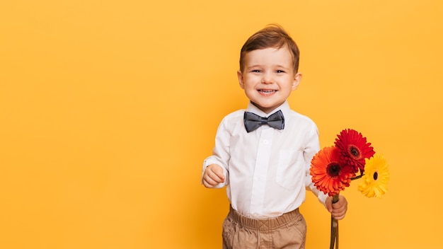 A boy in a white shirt, trousers and bow tie holds a bouquet of gerberas.